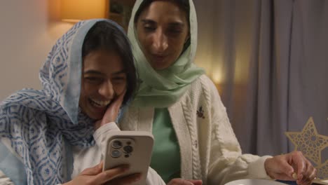 mother and daughter laughing at mobile phone sitting around muslim family table at home eating iftar meal breaking daily fast during ramadan 1