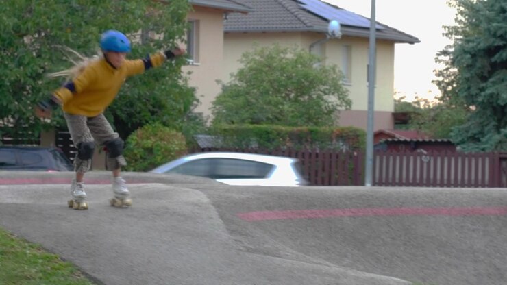 A wide static camera shot captures a young girl wearing a blue helmet roller skating along an asphalt pumptrack ridge near suburban houses in Kőbánya Sportliget park located in Budapest Hungary.