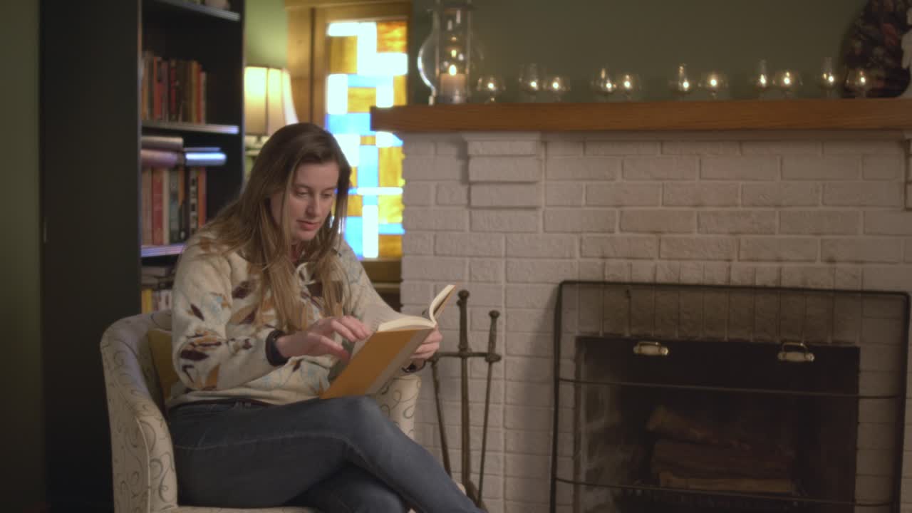 A woman in a sweater is quickly reading a book that she can't put down. Cozy fall setting in front of a fireplace. Shot in 60 frames