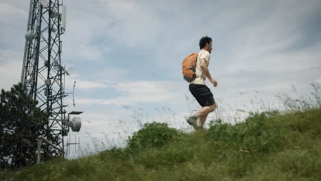 camera tracking a hiker with an orange backpack running up a hill past the radio tower on a rocky path surrounded with green summer grass