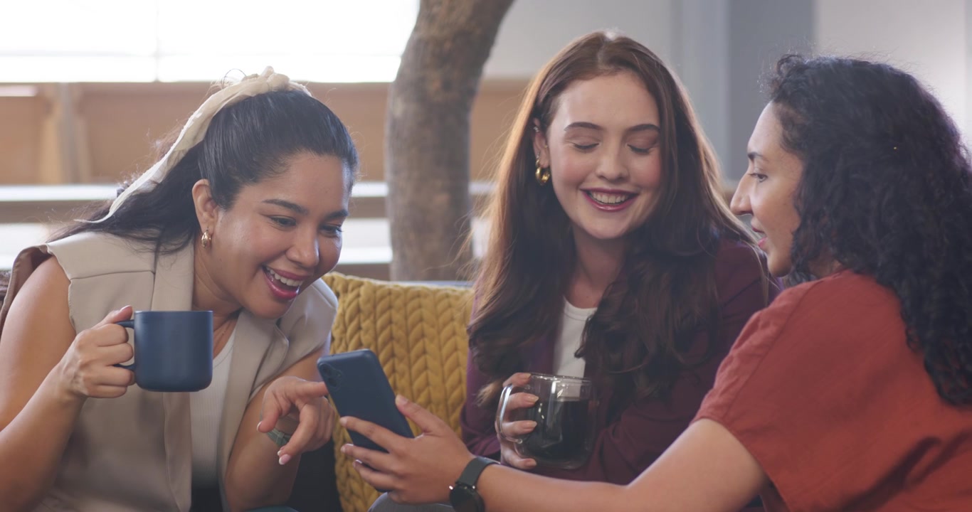 Three women sitting on couch, right woman presenting smartphone, sharing content while sipping mugs