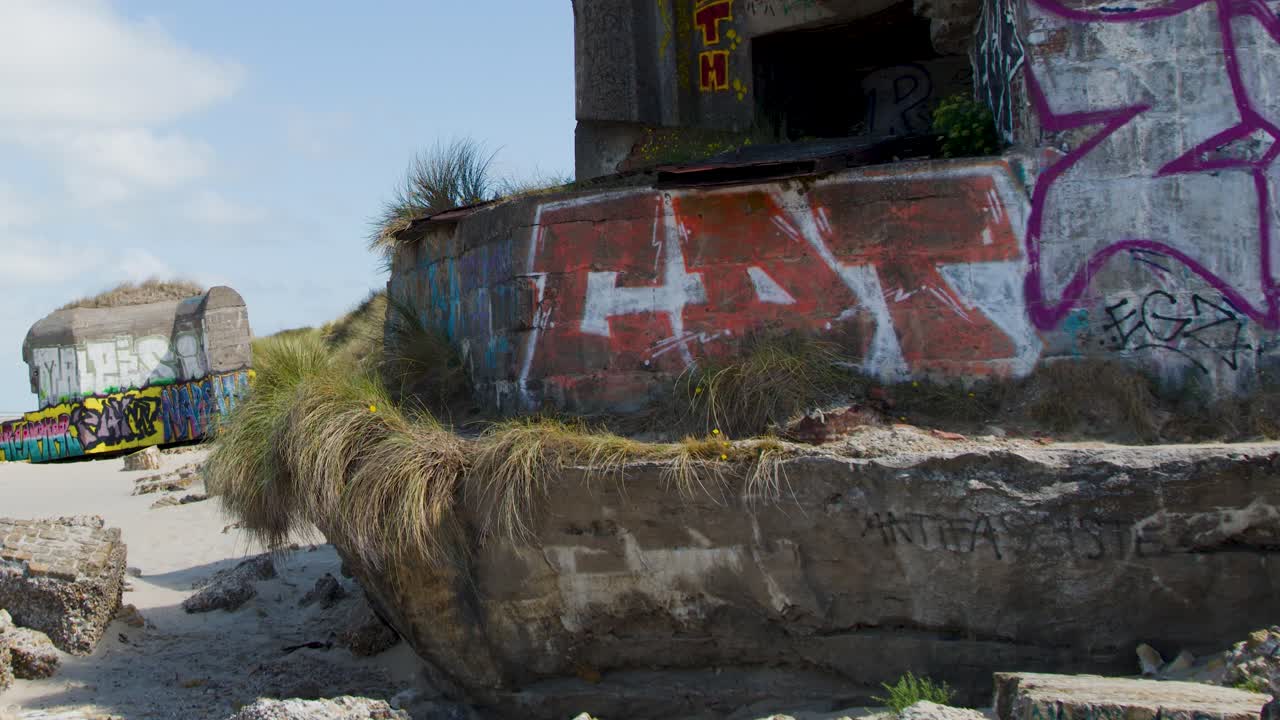 Daytime pan reveals historic concrete bunkers with graffiti, sand dunes, and ocean under clear sky