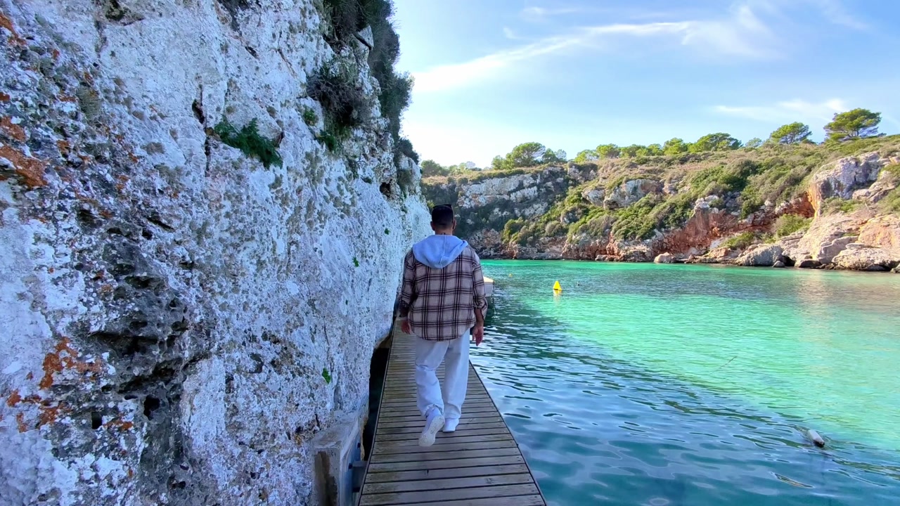 Man walking on wooden boardwalk along turquoise sea and limestone cliffs in Es Canutells, Menorca