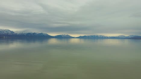 Flying-above-a-seemingly-endlessly-large-lake-with-seagulls-below-on-the-water-and-snowy-mountains-in-the-distance