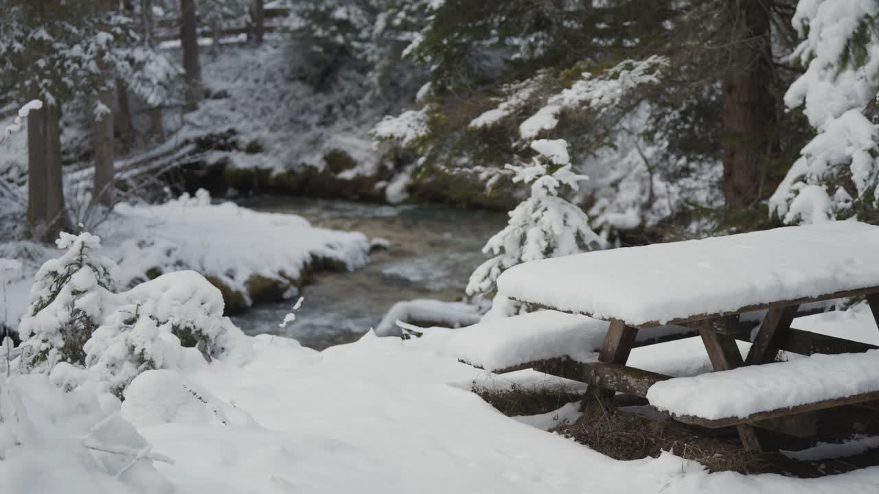 A snow-covered picnic table in the winter forest on the bank of the shallow mountain stream. Parallax video.