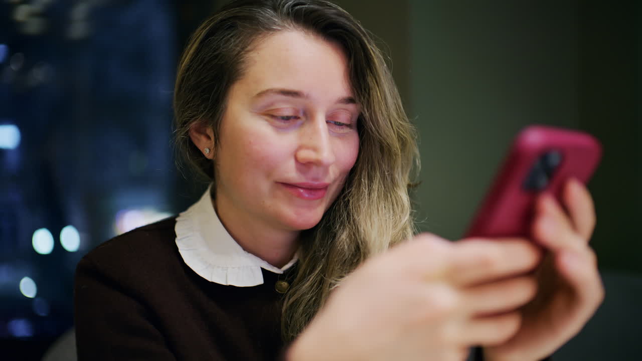 En kvinde indendørs om natten, blødt belyst, kigger på sin telefon med et let smil.