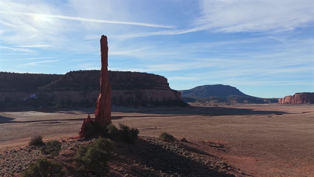 Un lento avance de dron cinematográfico se mueve detrás de un solitario pináculo de roca roja hacia magníficas mesetas desérticas, capturando la escarpada belleza de la arenisca y la escala dramática de una antigua vista de desierto árido