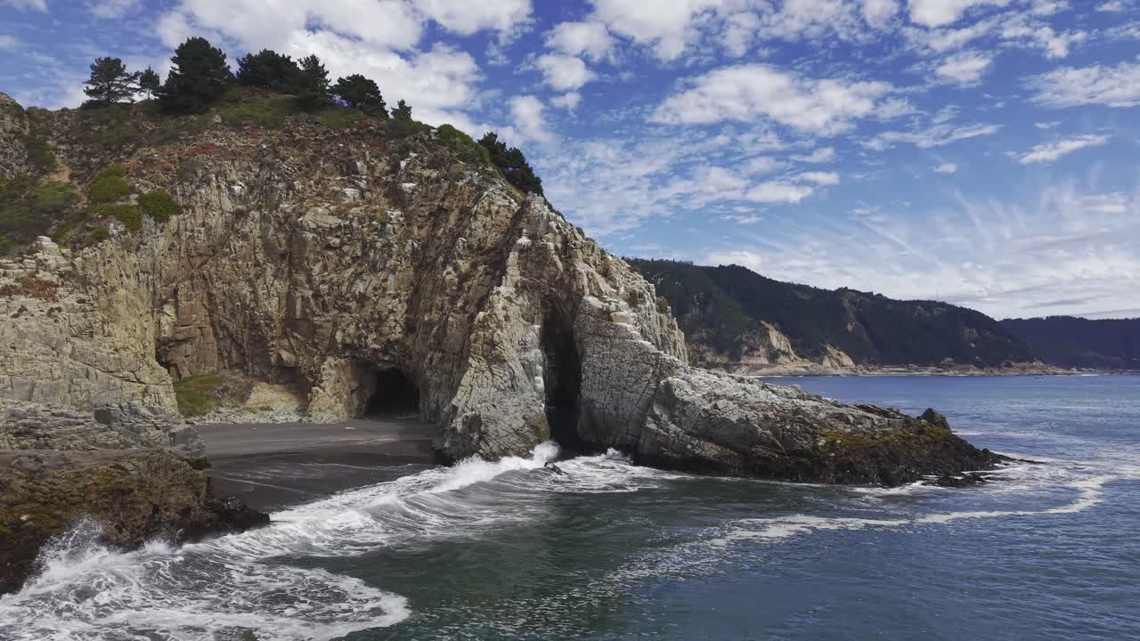 Scivolando velocemente verso una monumentale formazione rocciosa costiera con una grotta marina alla base della scogliera, mentre le onde lambiscono la cala di sabbia scura vicino a Constitución Maule Cile, 4K