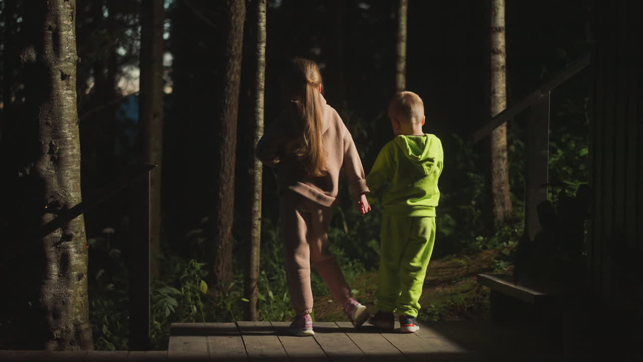 дети на площадке дома в диком летнем лесу. маленькая девочка с младшим братом стоит на гостиничном мешке у ночного леса. дети отдыхают в лесном лагере