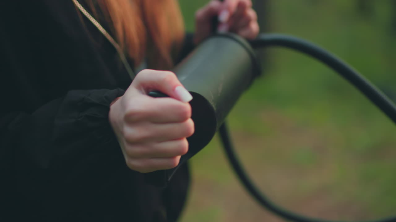 Close up of woman with long hair wearing black jacket with open zip while pumping nozzle using both hands, soft blur background of green forest visible behind hands and tool