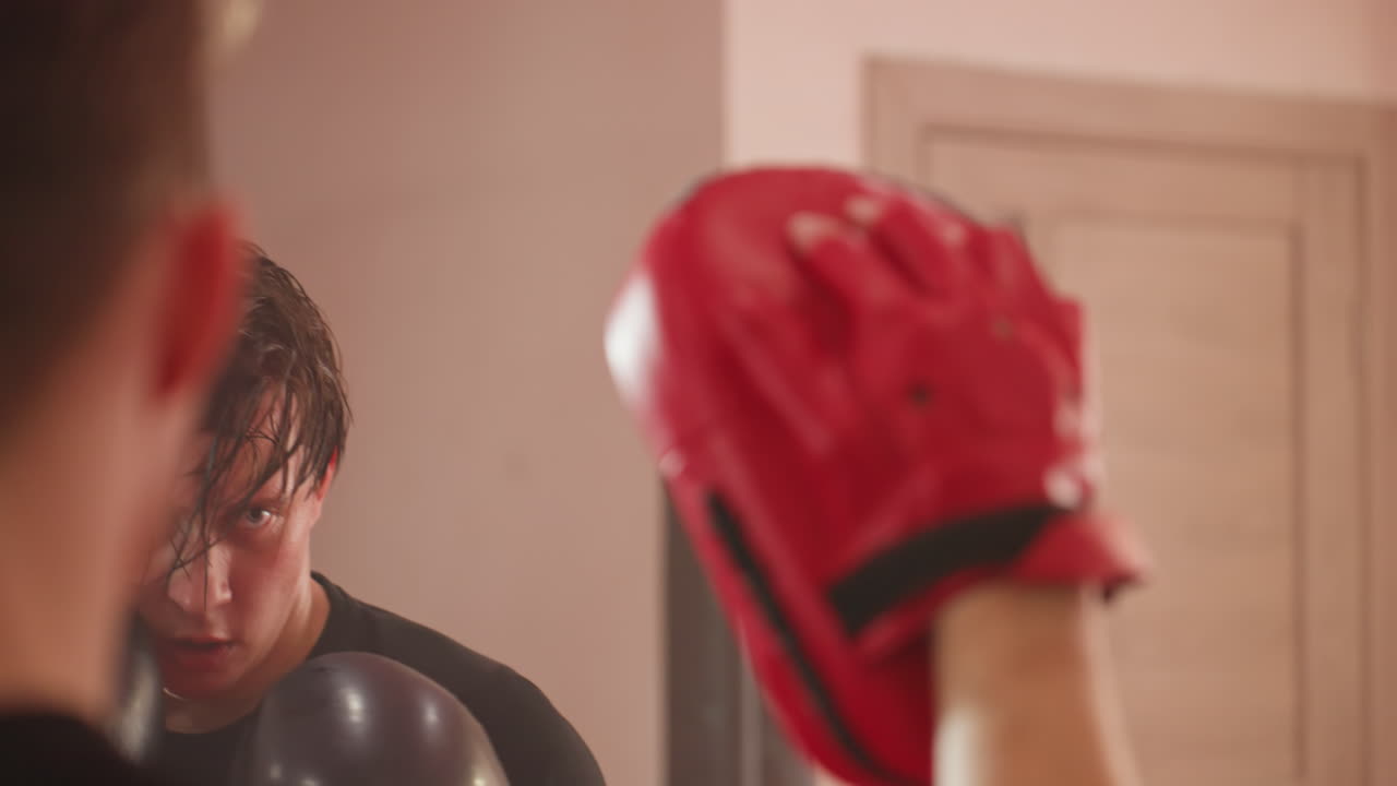 Focused boxer wearing black gloves trains with coach using red pads during intense sparring session, sweat dripping from face, eyes locked on target, demonstrating endurance, discipline in boxing gym
