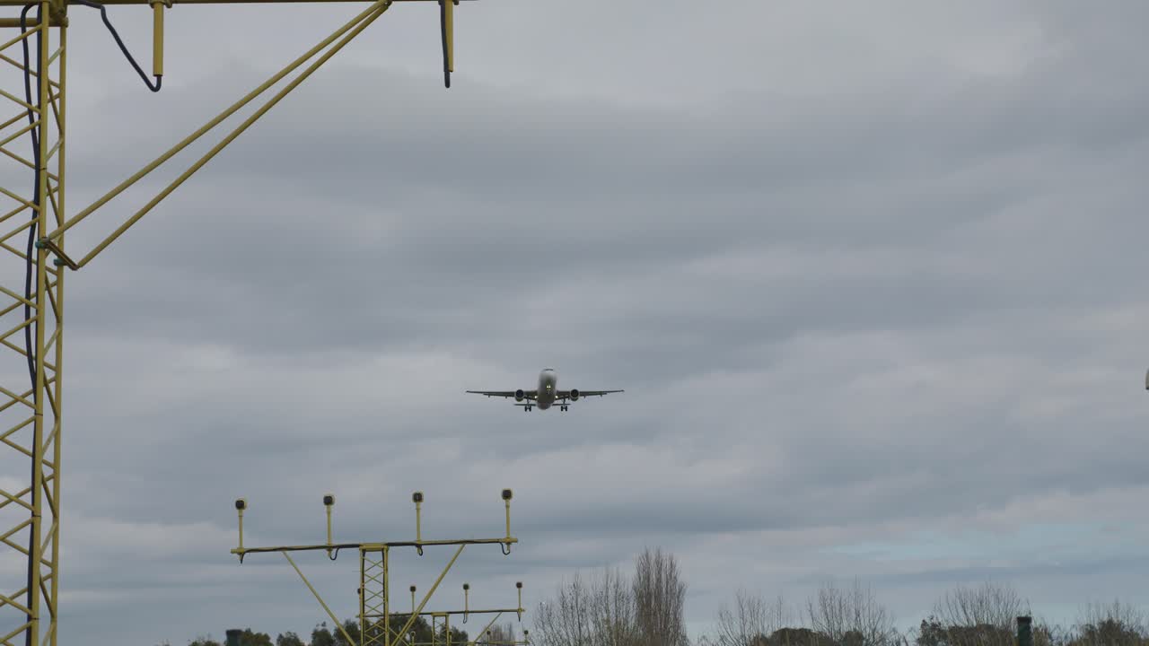 White passenger airplane approaching airport runway for landing, with landing gear extended and flaps down, against a backdrop of cloudy sky