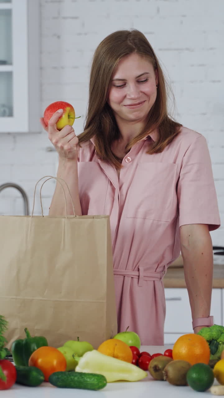 Lovely woman with fresh apple at home. Organic fruit and vegetables on the kitchen table and young female holding an apple. Healthy food. Vertical video
