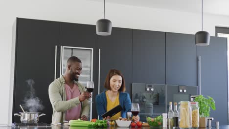 Video-of-happy-diverse-couple-preparing-food-making-a-toast-and-using-tablet-in-kitchen-at-home