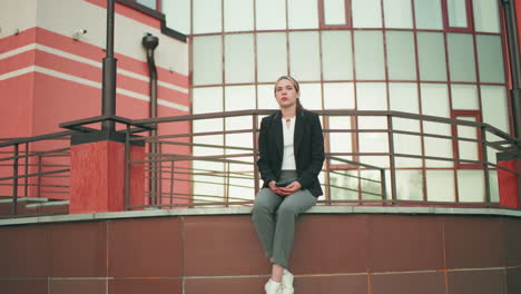 professional lady in black blazer seated on fence holding phone, modern building in background with urban design, natural lighting, legs crossed at ankle