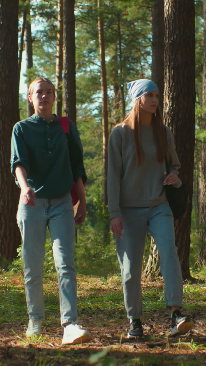 Two sisters walking through a sunlit forest, one wearing red backpack with cloth and the other with blue headscarf, as sunlight filters through trees and reflects gently on their faces