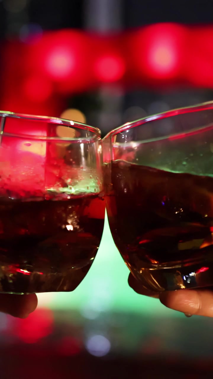 Two hands clink cocktail glasses filled with a dark beverage, set against colorful, out-of-focus bar lights. Shallow depth of field, festive mood