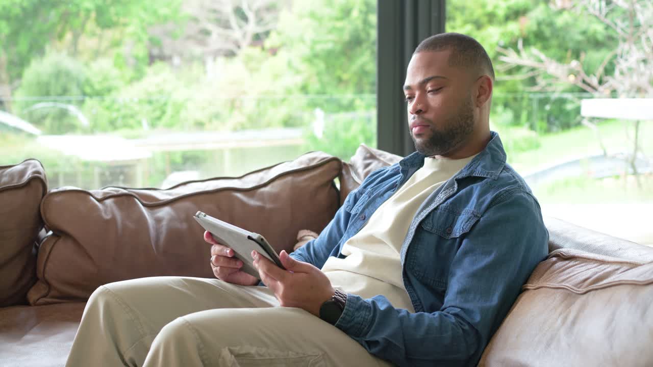 African American male swiping tablet on sofa by window, hearing sound, tilting head and laughing