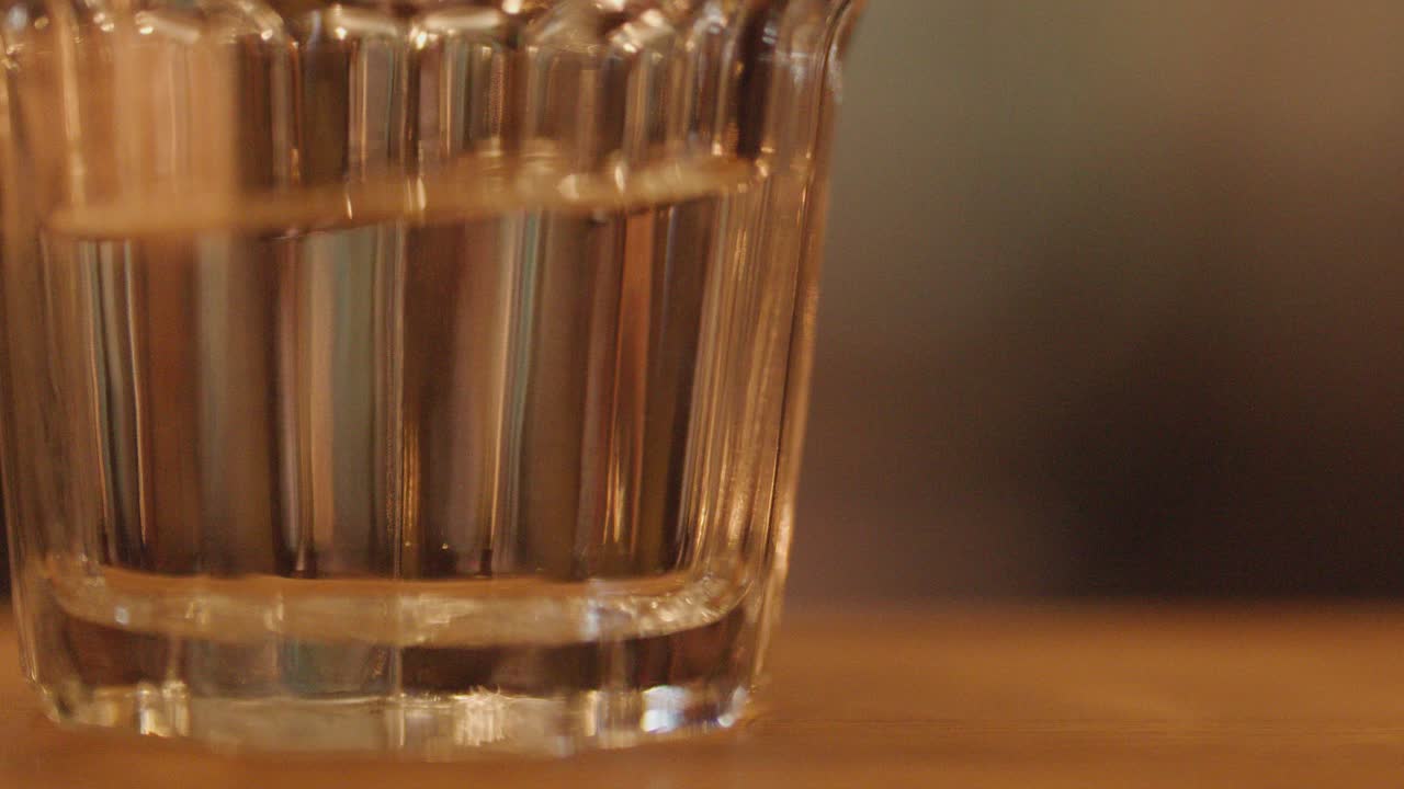 A hand places a clear glass of water on a wooden table in a softly lit restaurant at night, with a blurred background and warm bokeh lighting