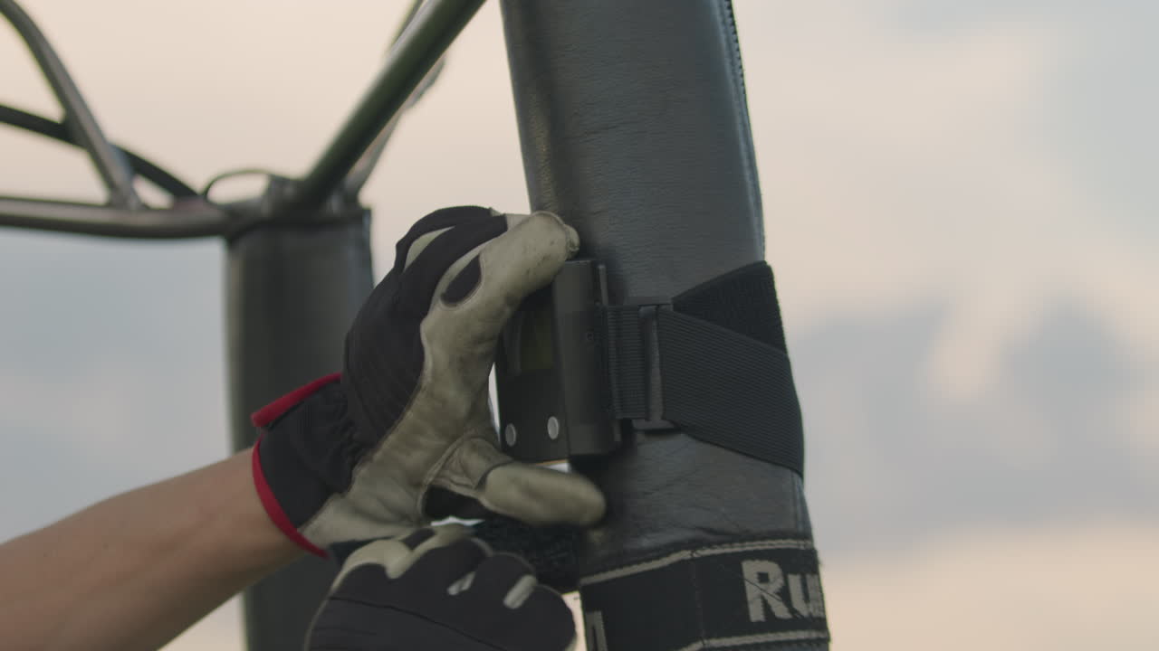 Close up of gloved hand adjusting black strap around vertical metal pole during outdoor equipment setup under cloudy sky, showcasing preparation process, and tool interaction in daylight
