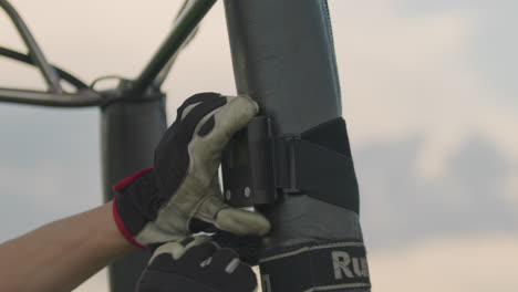 close up of gloved hand adjusting black strap around vertical metal pole during outdoor equipment setup under cloudy sky, showcasing preparation process, and tool interaction in daylight