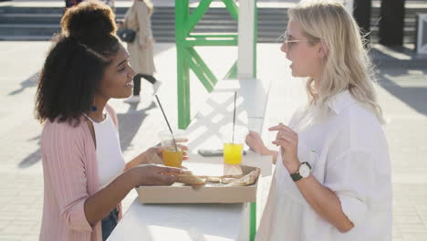 female friends chatting and laughing while sharing pizza and drinking, standing at an outdoor table in the street 2