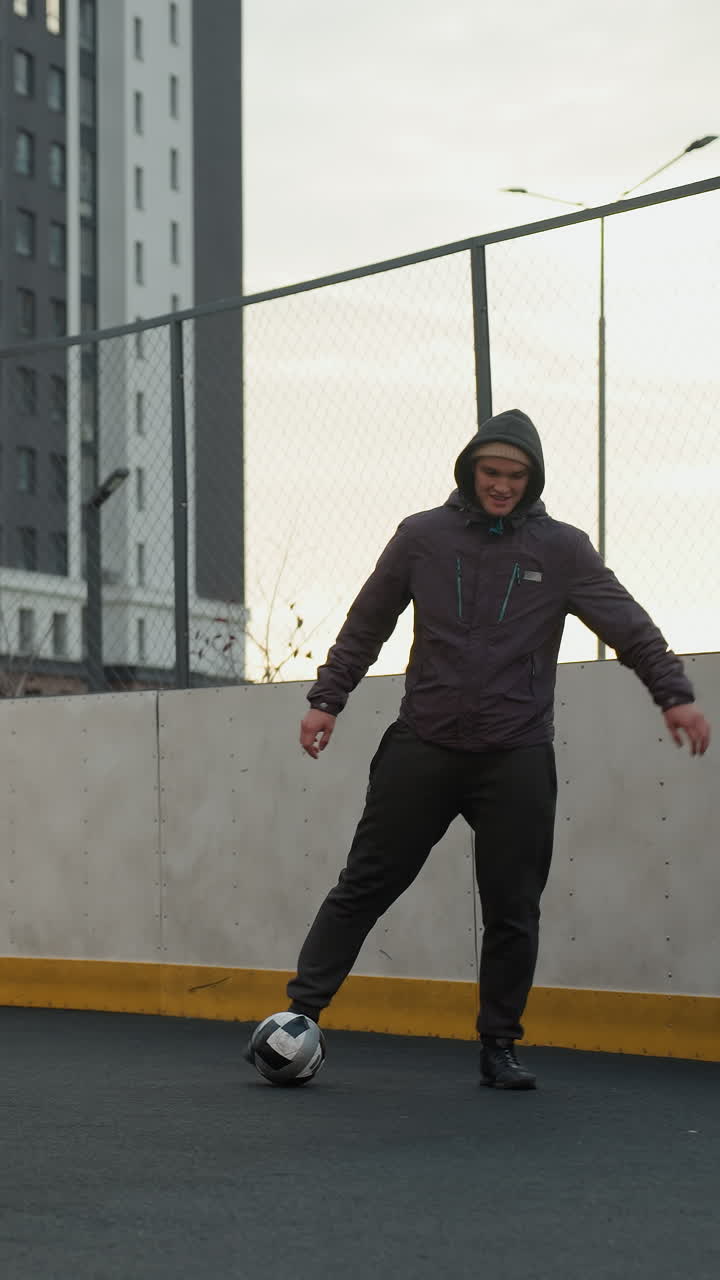 entrenador hábilmente driblando la pelota de fútbol en la arena deportiva durante una sesión de entrenamiento, mostrando control preciso y trabajo de pies, el fondo presenta edificios urbanos modernos, esgrima y luz solar suave