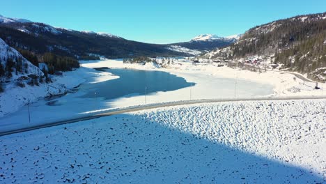 Langsame-Luftaufnahme-Des-Von-Statkraft-Betriebenen-Tunhovd-Staudamms-Mit-Dem-Tunhovdfjord-Wasserreservoir-Im-Inneren-–-Niedriger-Füllgrad-An-Sonnigen-Wintertagen-–-Straßenkreuzung-Oben-Mit-Blauem-Himmelshintergrund-In-Nore-Und-Uvdal