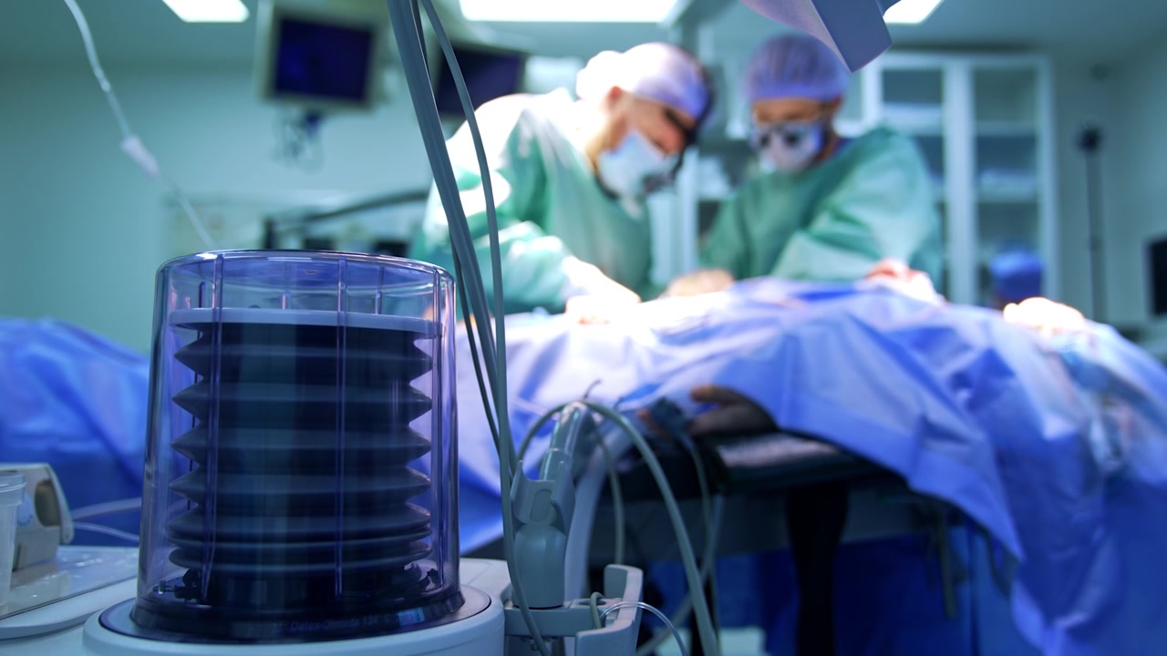 Artificial lung ventilator working in surgery room. Two surgeons operate a patient at backdrop in blur.