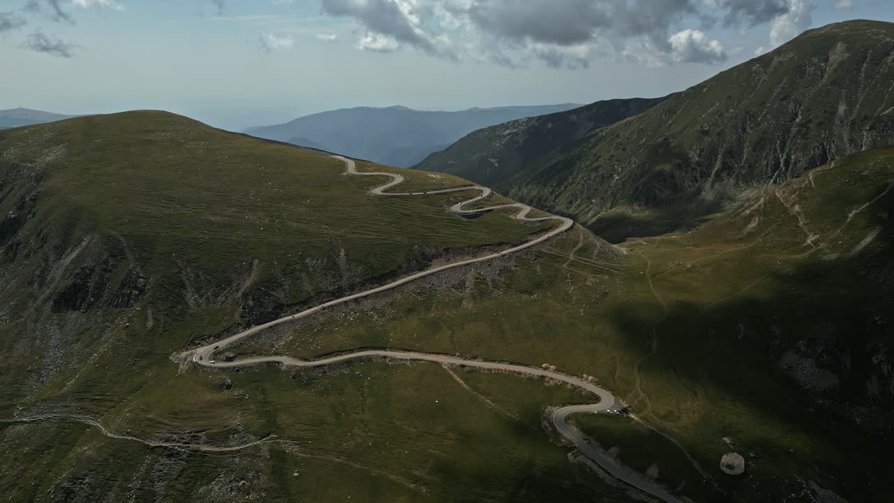 Stunning vista of the winding roads in Romania's Transalpina, set against lush greenery, rugged hills, and a vast horizon beneath a cloud-dappled sky