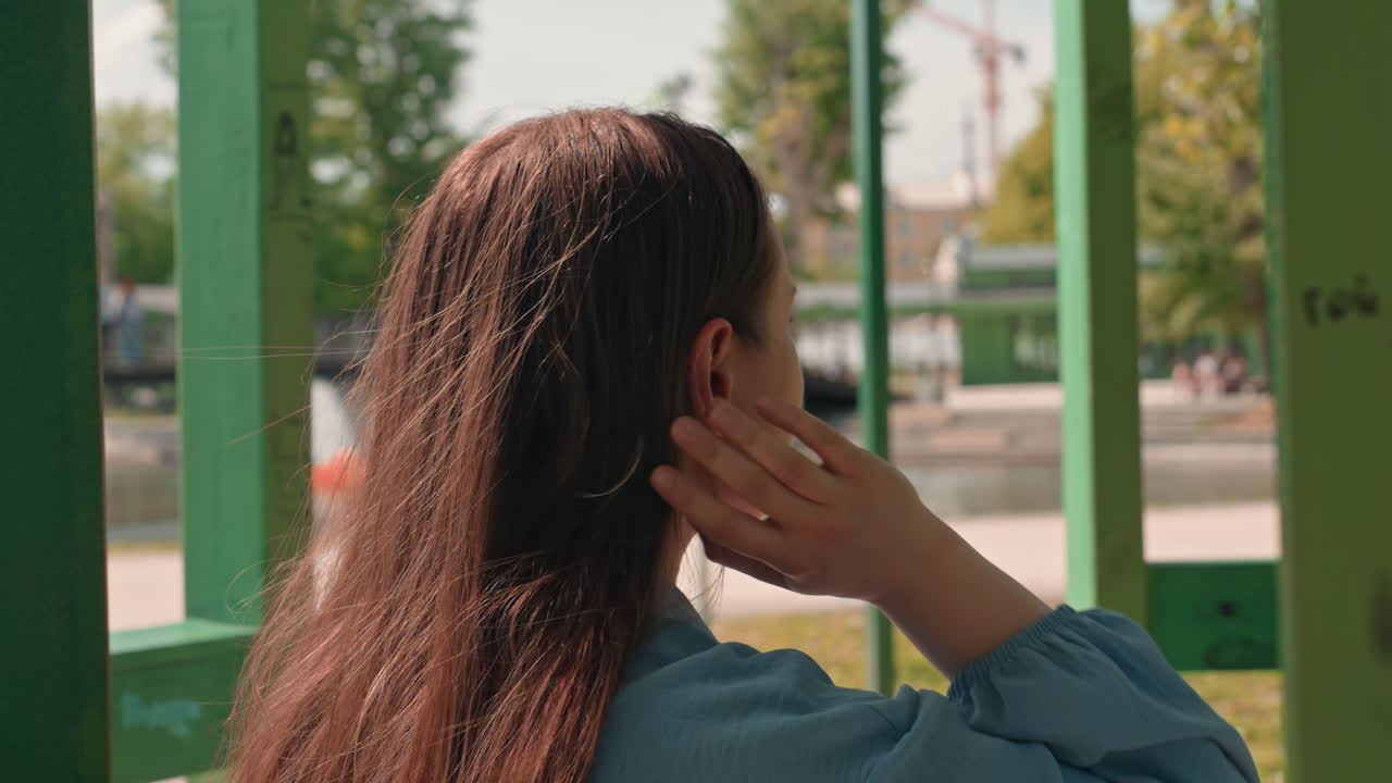 여성이 머리카락을 만지고, 도시 풍경 속에서 평범한 여성의 모습이 보여요. 백인 여성이 도시 공원 배경으로 머리카락을 정리하고, 낙서 벽 옆에서 편안한 복장으로 머리카락을 만지며 차분하게 서 있어요.