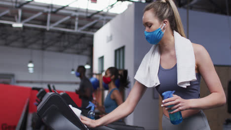 Mujer-Caucásica-En-Forma-Con-Mascarilla-Limpiando-La-Cinta-De-Correr-Con-Desinfectante-En-El-Gimnasio