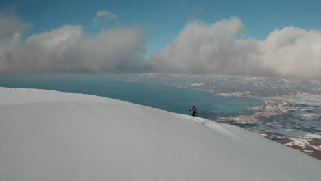 Splitboarder-on-a-snowy-Hokkaido-mountain-with-Niseko-resort-in-the-distance-under-a-bright-blue-sky