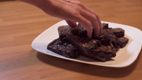 Mano-De-Mujer-Agarrando-Un-Brownie-De-Una-Pila-De-Brownies-En-Un-Plato-Blanco-Colocado-En-Una-Mesa-De-Comedor-De-Madera-Mientras-La-Cámara-Se-Desliza-Hacia-La-Derecha