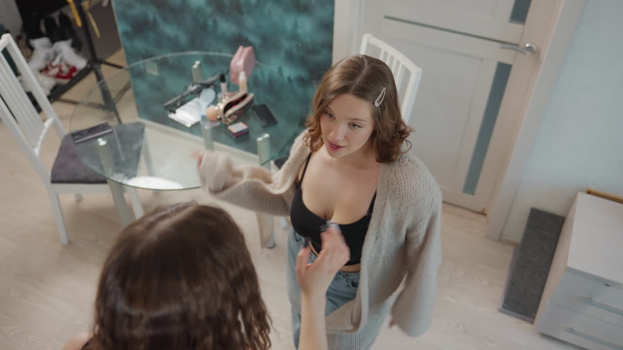 Young Woman Greets Friend In Bedroom With OpenArms Hug, Playful Kiss And Laughter, Casual Outfits, Makeup And Mirror On Glass Table, Cozy Modern Interior, Natural Lighting, Candid Warm Atmosphere