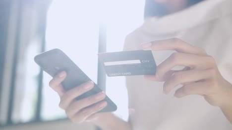 beautiful woman customer holding credit card and smartphone sitting in front of window at home. she using instant easy mobile payments making purchase in online store. e-banking app. 4k resolution.