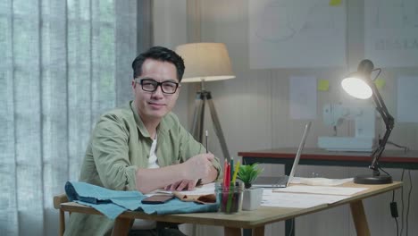 직물, 노트북, 스마트폰을 가진 아시아인 디자이너가 사무실에서 레이아웃 본드를 그리는 동안 카메라를 향해 미소 짓고 있습니다.