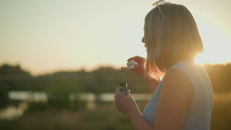 mujer con una camiseta sin mangas azul con gafas de sol en la cabeza soplando burbujas durante la hora dorada, la luz del sol iluminando su cabello, con un fondo natural borroso