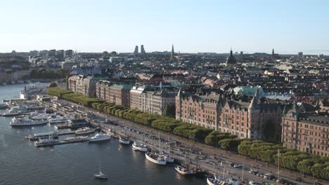 traffic, pedestrians and boats in dock on strandvägen and strandkajen, stockholm during sunny evening with old apartment buildings and skyline visible