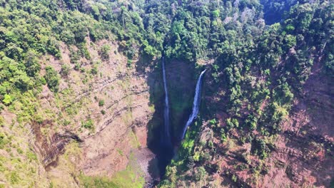 twin waterfalls carve through dense forest as they descend a steep cliff into a shadowed gorge in pakse, laos. the dramatic terrain and vibrant canopy create a striking natural contrast from above