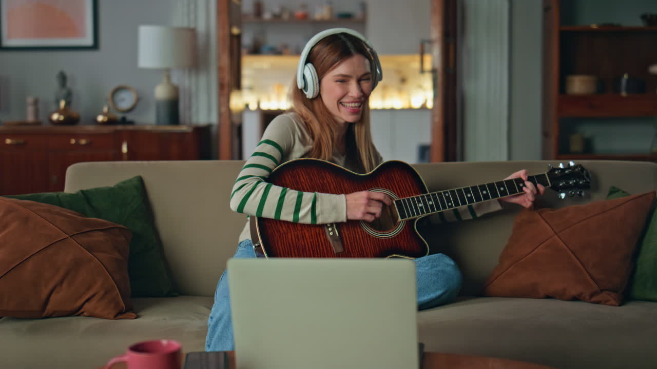 Smiling woman playing guitar in headphones at living room couch. Happy guitarist