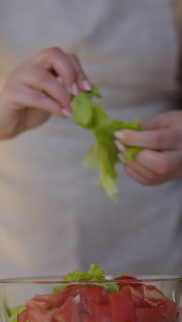 concentrated young woman in apron tears juicy lettuce leaves adding to delicious salad at table in light kitchen close view slow motion