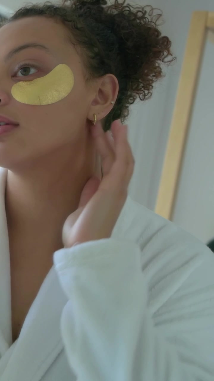 Vertical video: Grabbing gold patches, woman in robe smoothing them at vanity for under-eye care