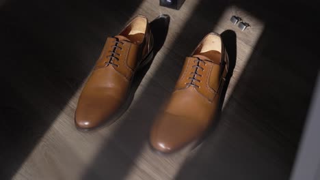 brown leather wedding shoes placed on wooden floor, sunlight casting shadows, cufflinks nearby