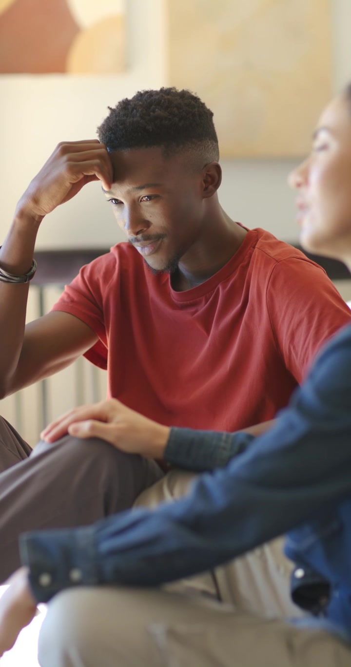 Vertical video: Diverse couple man rubbing forehead while talking, woman reaching, soothing on sofa