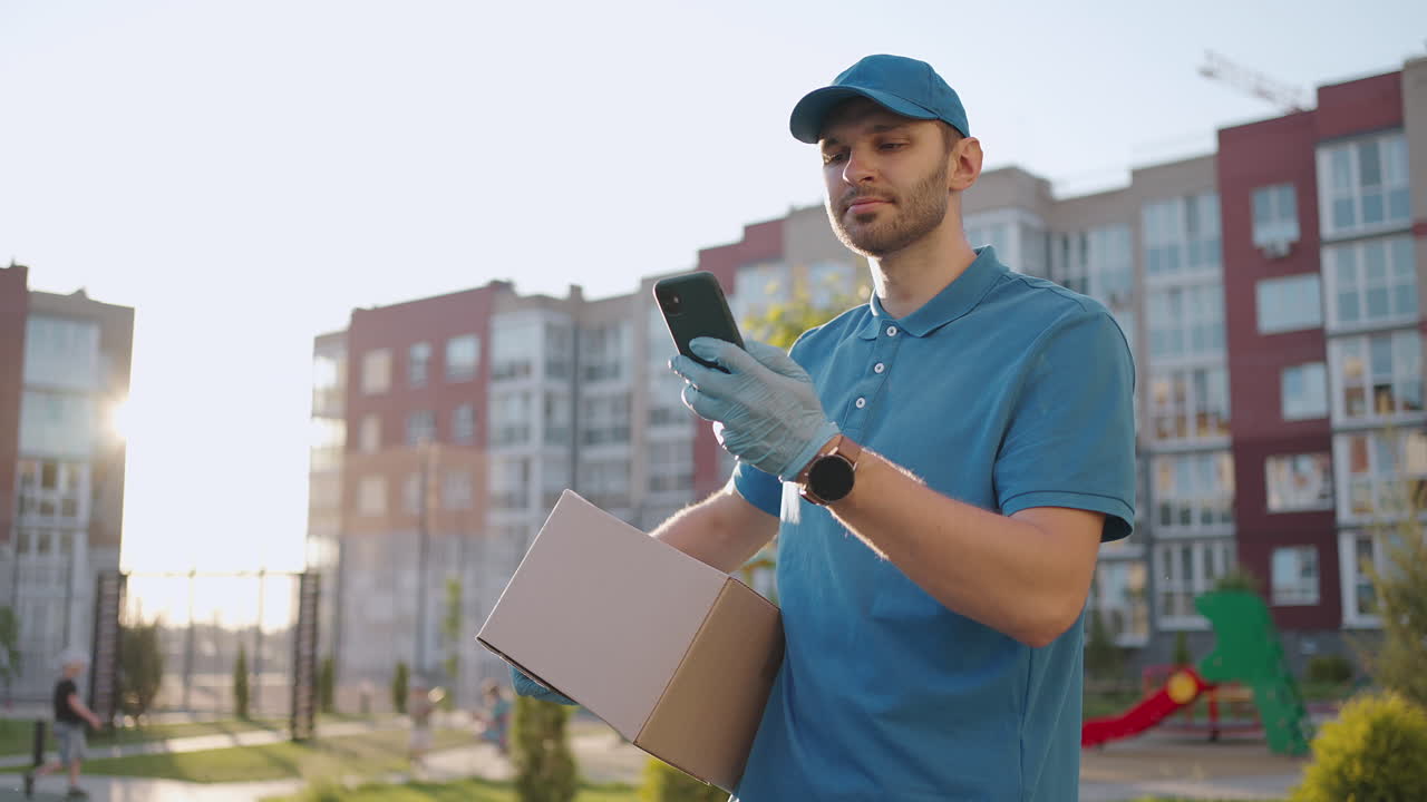 el cartero con gafas lleva el paquete y mira la dirección de entrega a través de un teléfono móvil. busque la dirección del cliente de entrega. el tipo de entrega con una tapa y una caja