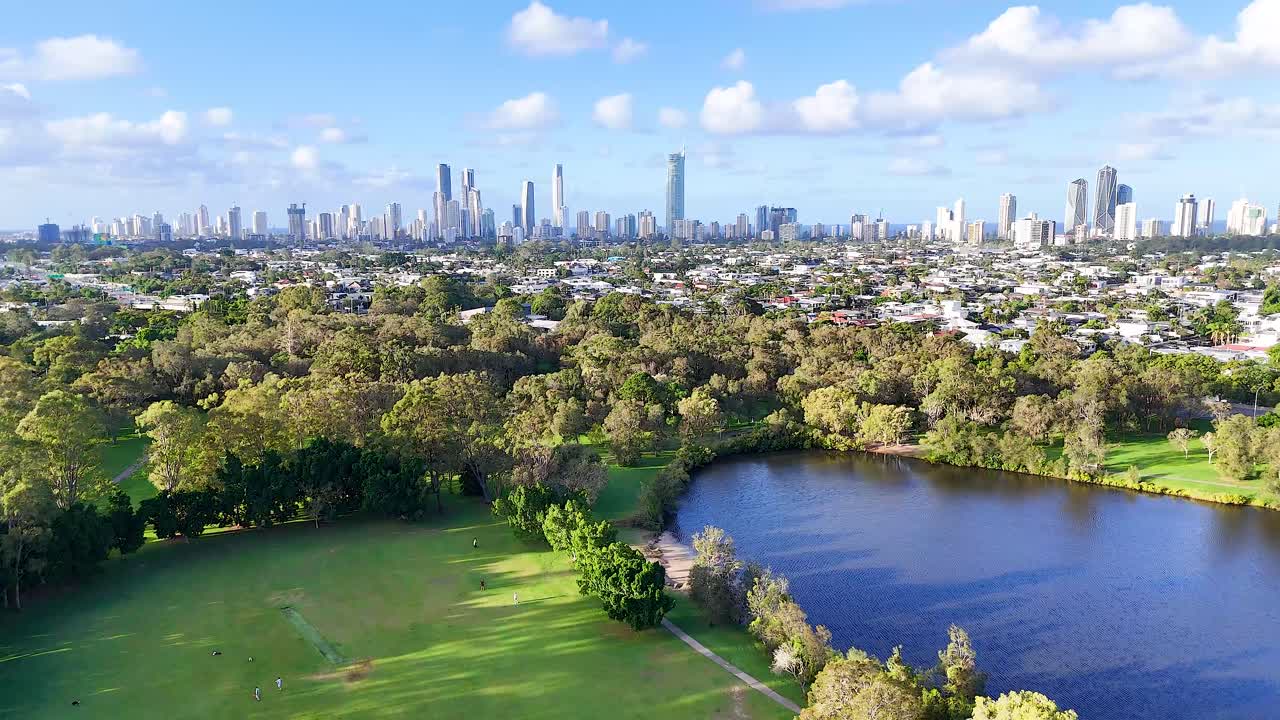Aerial footage captures Gold Coast's skyline, lush park, and serene water body under bright daylight