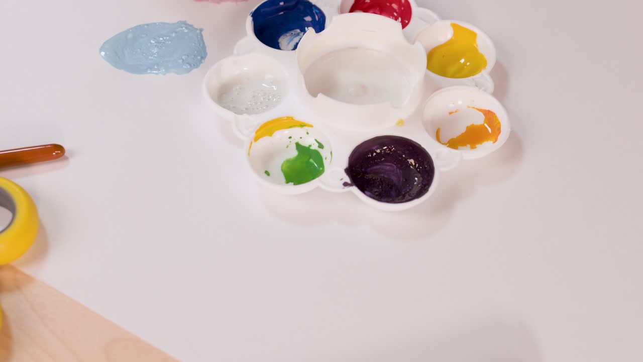 Artist hand mixes watercolor paints on palette, creating new color on white paper, top view