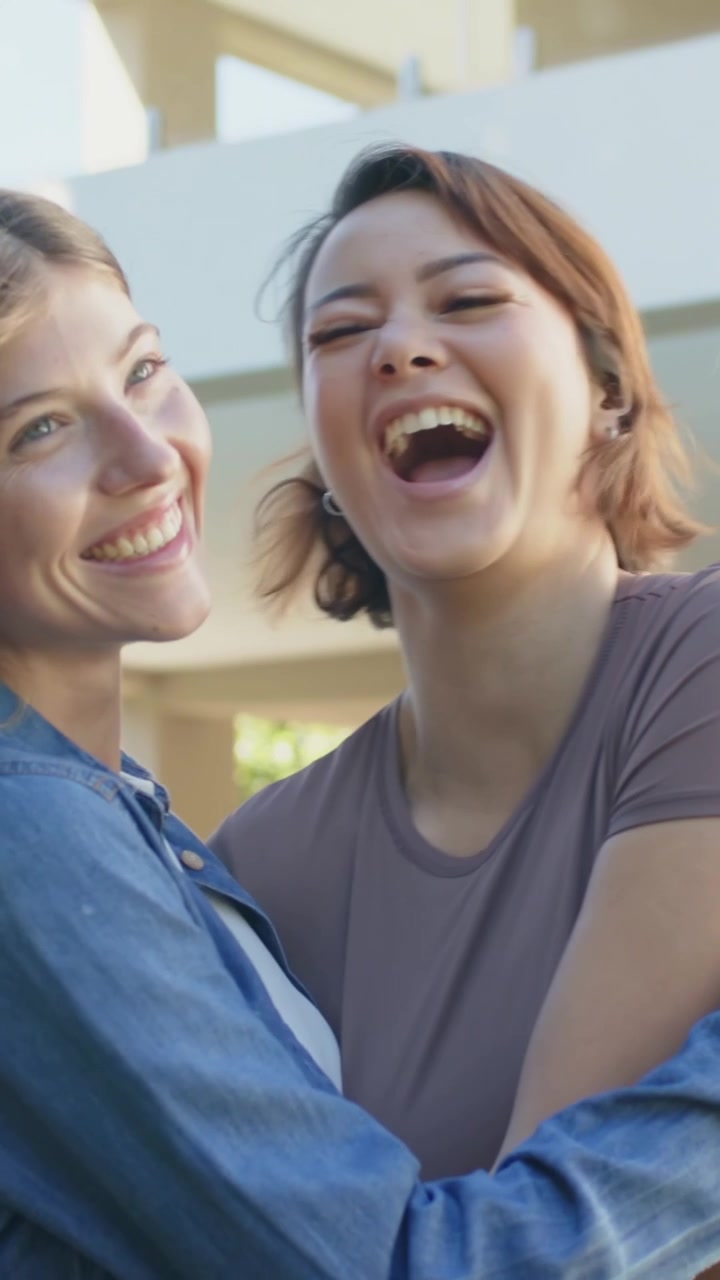 Vertical video: One woman leaning, two women hugging and laughing on patio, denim shirt mauve tee