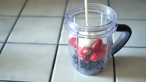 pouring milk yogurt frozen blueberries and strawberries into blender to make smoothie or fitness shake